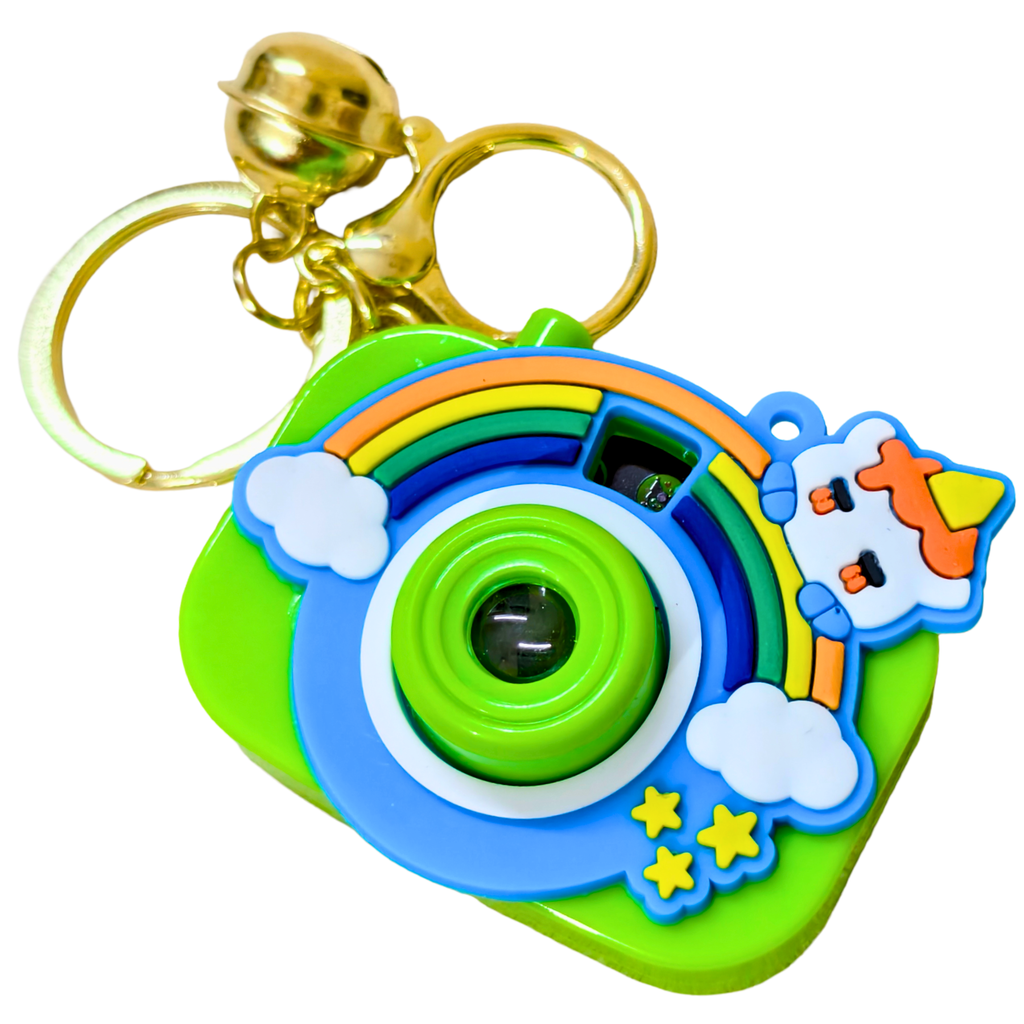 A colorful toy camera keychain with a gold bell and split ring. The camera body is bright green with a blue lens area, decorated with a rainbow, white clouds, yellow stars, and a cartoon character peeking from behind on the right.