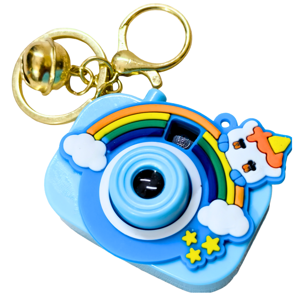 A light blue camera-shaped keychain with a gold clasp and bell, decorated with a rainbow, white clouds, yellow stars, and a small cartoon character on a white background.