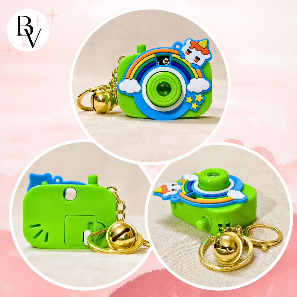 Three circular product views of a small camera-shaped keychain charm in bright green, decorated with a rainbow, clouds, stars, a unicorn, and a gold bell keyring, shown on a soft pink background.