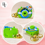 Three circular product views of a small camera-shaped keychain charm in bright green, decorated with a rainbow, clouds, stars, a unicorn, and a gold bell keyring, shown on a soft pink background.