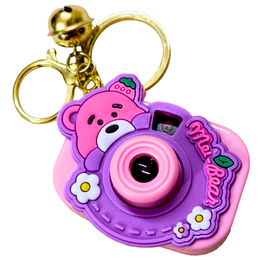 A pink and purple camera-shaped keychain with a cartoon bear face, small white flowers, and a gold bell keyring on a plain white background.