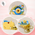 A small yellow camera-shaped keychain charm shown in three circular close-up views on a soft pink background. The charm has a rainbow, clouds, stars, a small animal character, gold-tone key rings, and a small bell.