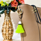 A close-up of a tan pebbled leather handbag with a bright neon yellow-green shuttlecock keychain hanging from the zipper. The handbag is shown next to a decorative textured vase with flowers.