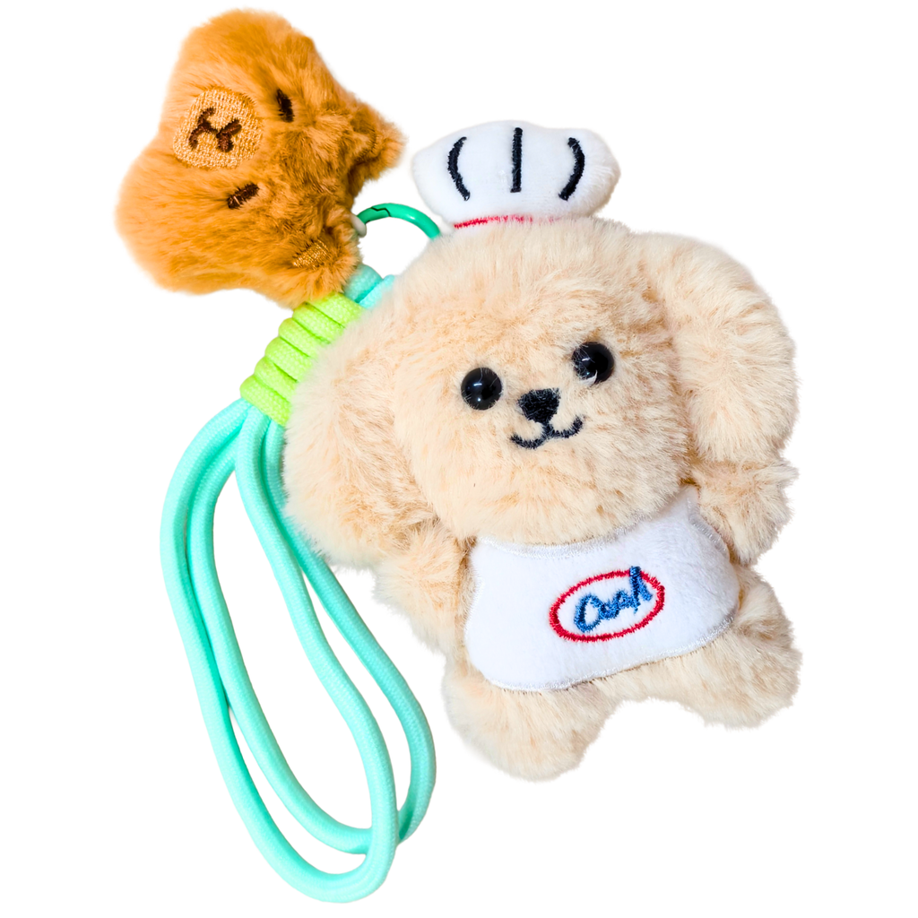 A plush dog keychain or car accessory featuring a cream-colored teddy bear wearing a white chef hat and bib with the word 'Cook' embroidered in blue and red. A small orange plush face hangs from a green ring at the top, and a mint green loop extends from the side.