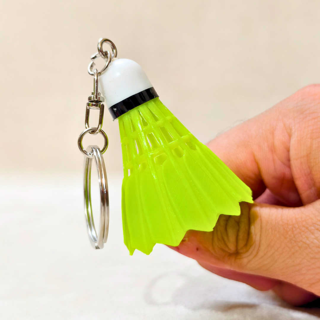 A small plastic shuttlecock-shaped keychain in bright neon yellow-green with a white top, black band, and silver metal key ring, held by a hand against a light beige background.