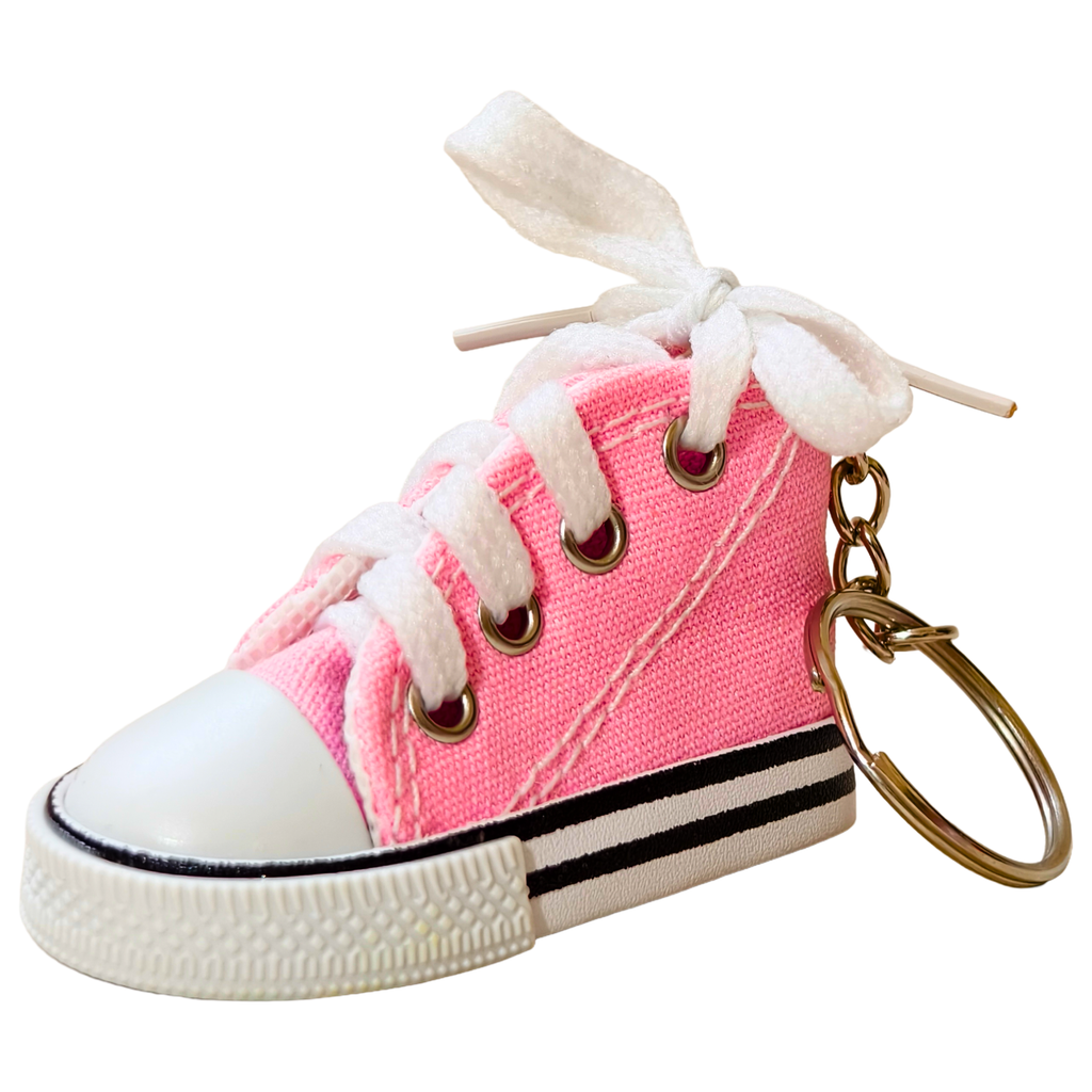 A miniature pink canvas high-top sneaker keychain with white laces tied in a bow, a white rubber toe cap, black and white stripes on the sole, and a gold metal key ring.
