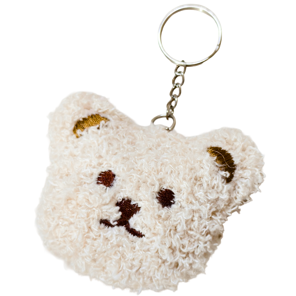 A small plush bear-face keychain . The keychain is cream-colored with embroidered brown facial features and gold-toned ear accents, attached to a silver metal ring and chain on a white background.