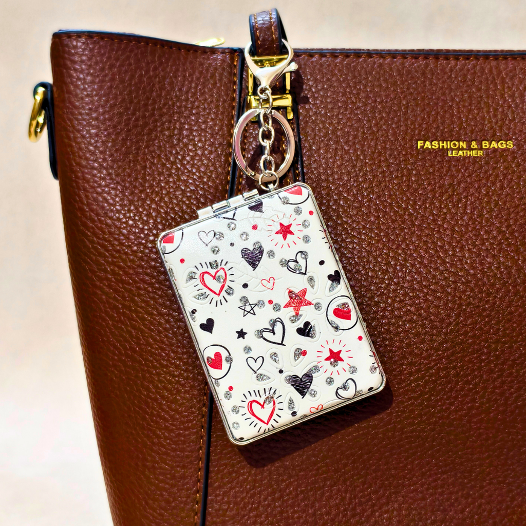 A close-up of a brown textured leather tote bag with a gold-tone zipper and a decorative keychain charm hanging from it. The charm has a white background with a pattern of red, black, and gray hearts, stars, and sunburst motifs.