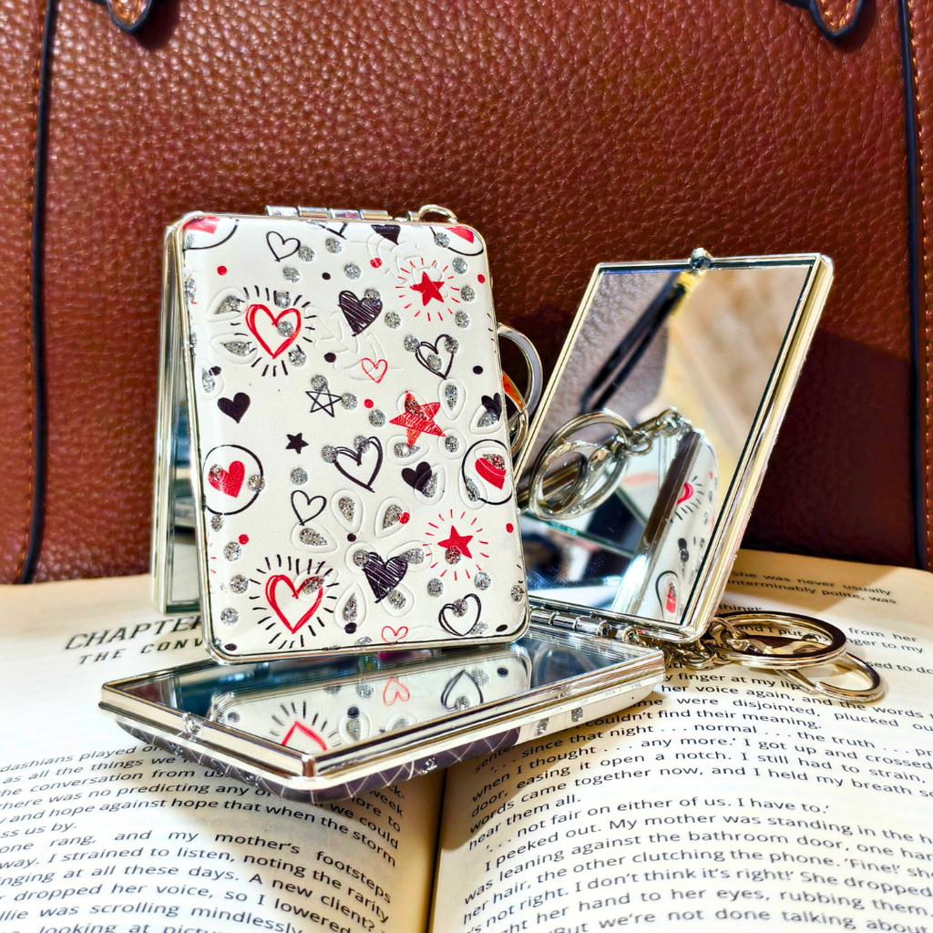 A small silver metal folding compact mirror with a white cover decorated with red, black, and gray heart and star illustrations, displayed on top of an open book against a brown leather background.