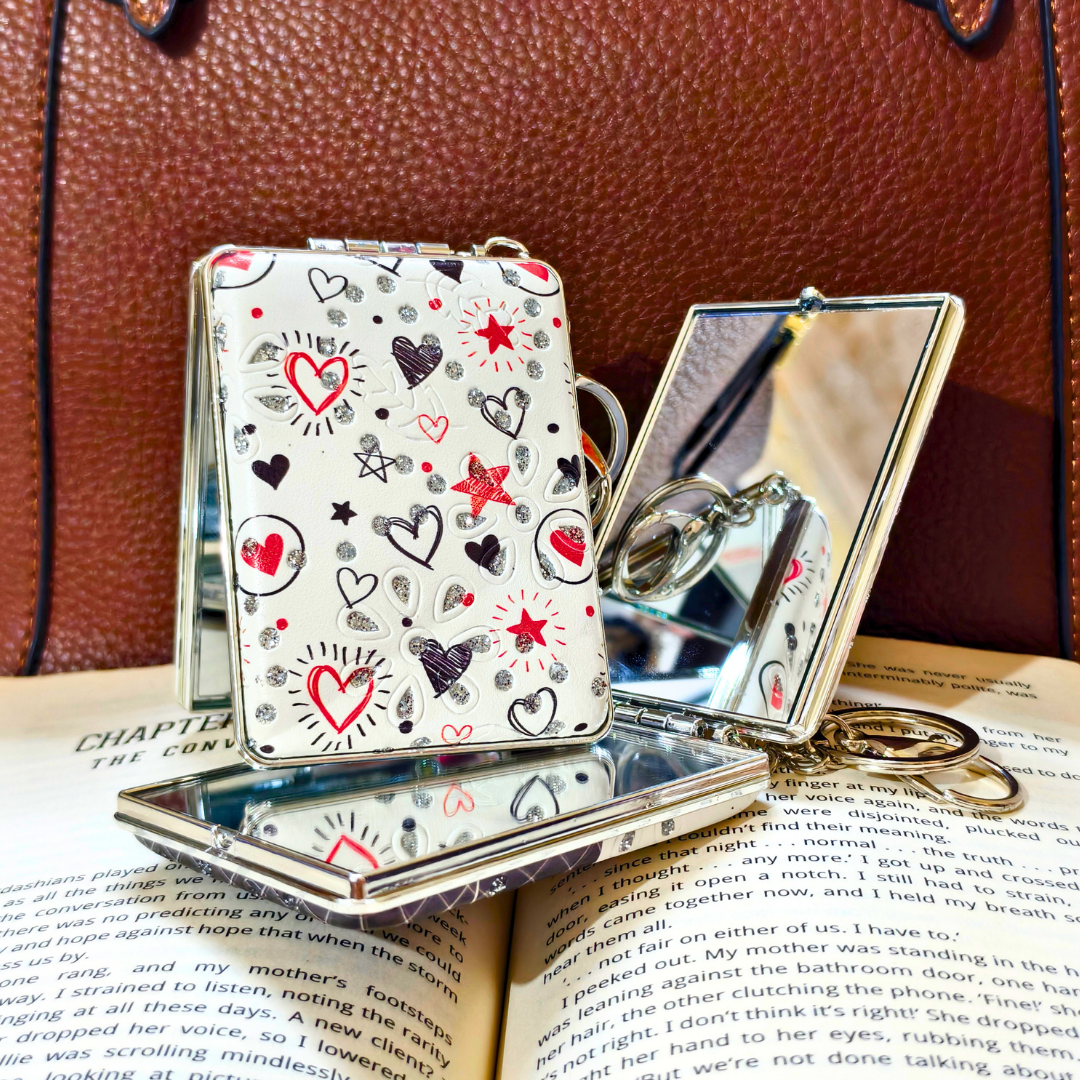 A small silver metal folding compact mirror with a white cover decorated with red, black, and gray heart and star illustrations, displayed on top of an open book against a brown leather background.