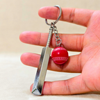 silver metal Cricket Bat and Ball  keychain with a small Cricket Ball attached, hold in hand  against a plain beige background.


