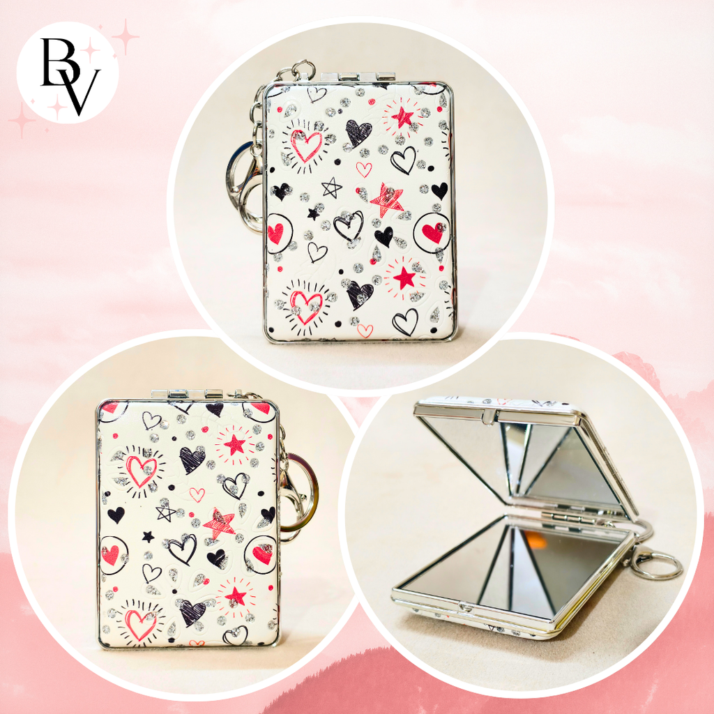 A compact folding mirror with a white background decorated with red, black, and pink heart and star doodles, shown in three circular views: closed, open, and open with the mirror flipped up. A 'BV' logo is visible in the top left corner.