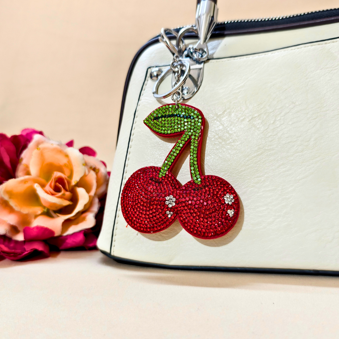 Red Shiny Cherry keychain with a metal latch placed on a white purse beside a flower in front of a beige background
