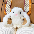 A white fluffy plush bunny toy with black eyes and a small pink nose, resting on an open book in front of a brown leather surface with a patterned scarf.