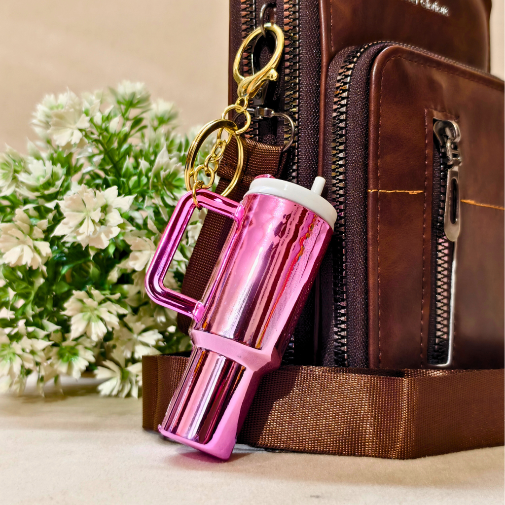 A shiny metallic pink travel tumbler with a handle and gold keychain rings, shown resting against a brown leather bag with white flowers in the background.