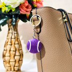 A purple tennis ball keychain charm with white curved stripes hangs from a silver metal key ring attached to a tan textured handbag. A decorative vase with flowers sits in the background.