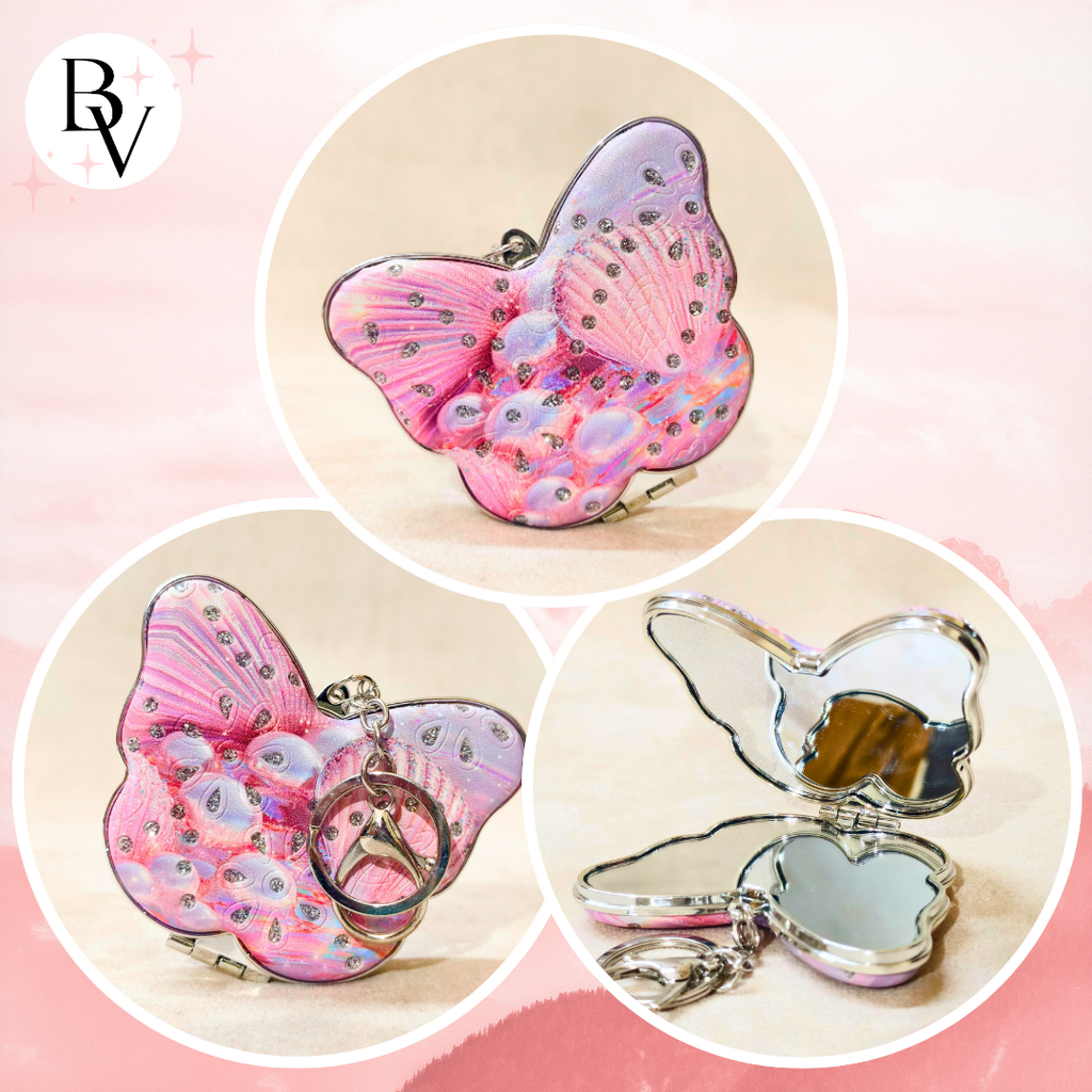 A butterfly-shaped compact mirror with a pink and lavender iridescent wing design, rhinestone embellishments, and a silver-tone metal frame. The collage shows the closed compact, the compact with a key ring attached, and the compact open to reveal a mirror inside, set against a soft pink background.