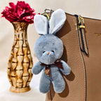 Plush bunny keychain attached to a brown leather bag with a wooden vase and red flowers in the background.