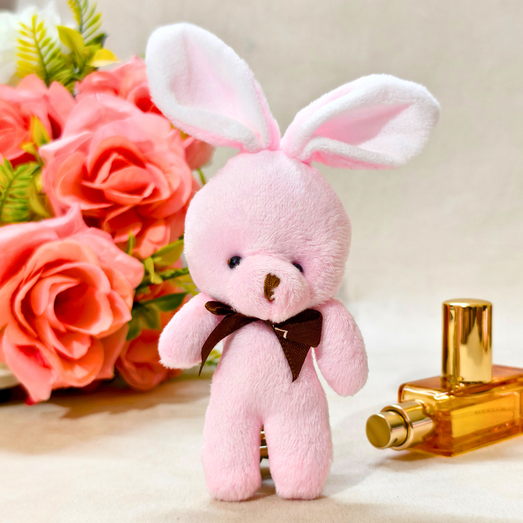A pink plush bunny toy with long floppy ears, a dark brown bow tie, and a small stitched nose, posed in front of pink roses and a gold perfume bottle on a light beige surface.
