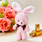 A pink plush bunny toy with long floppy ears, a dark brown bow tie, and a small stitched nose, posed in front of pink roses and a gold perfume bottle on a light beige surface.