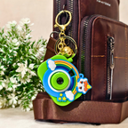 A colorful silicone camera-shaped keychain charm with a gold key ring and bell, shown hanging from a brown leather bag next to small white flowers.