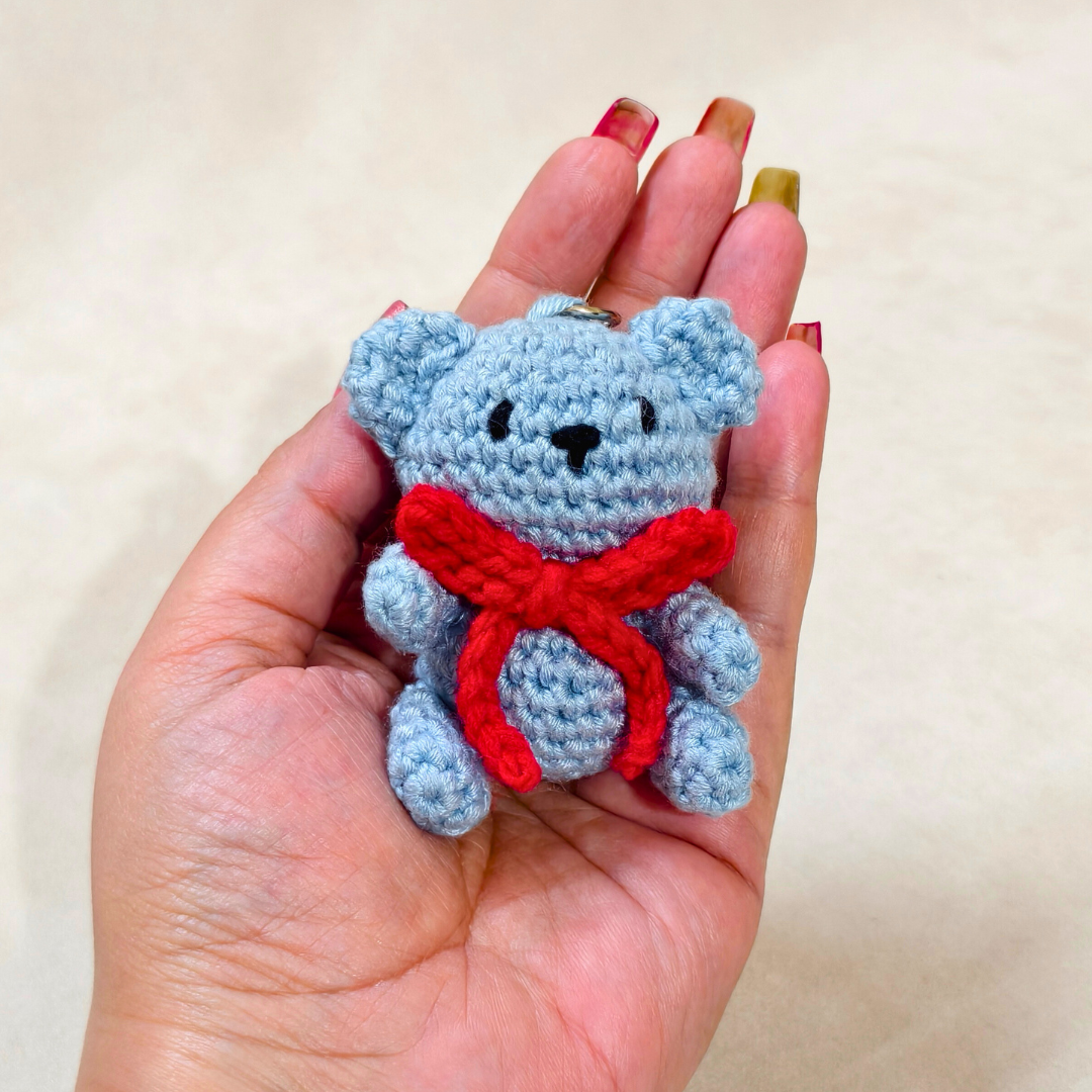 Hand holding a small blue teddy bear with a red bow tie against a plain background
