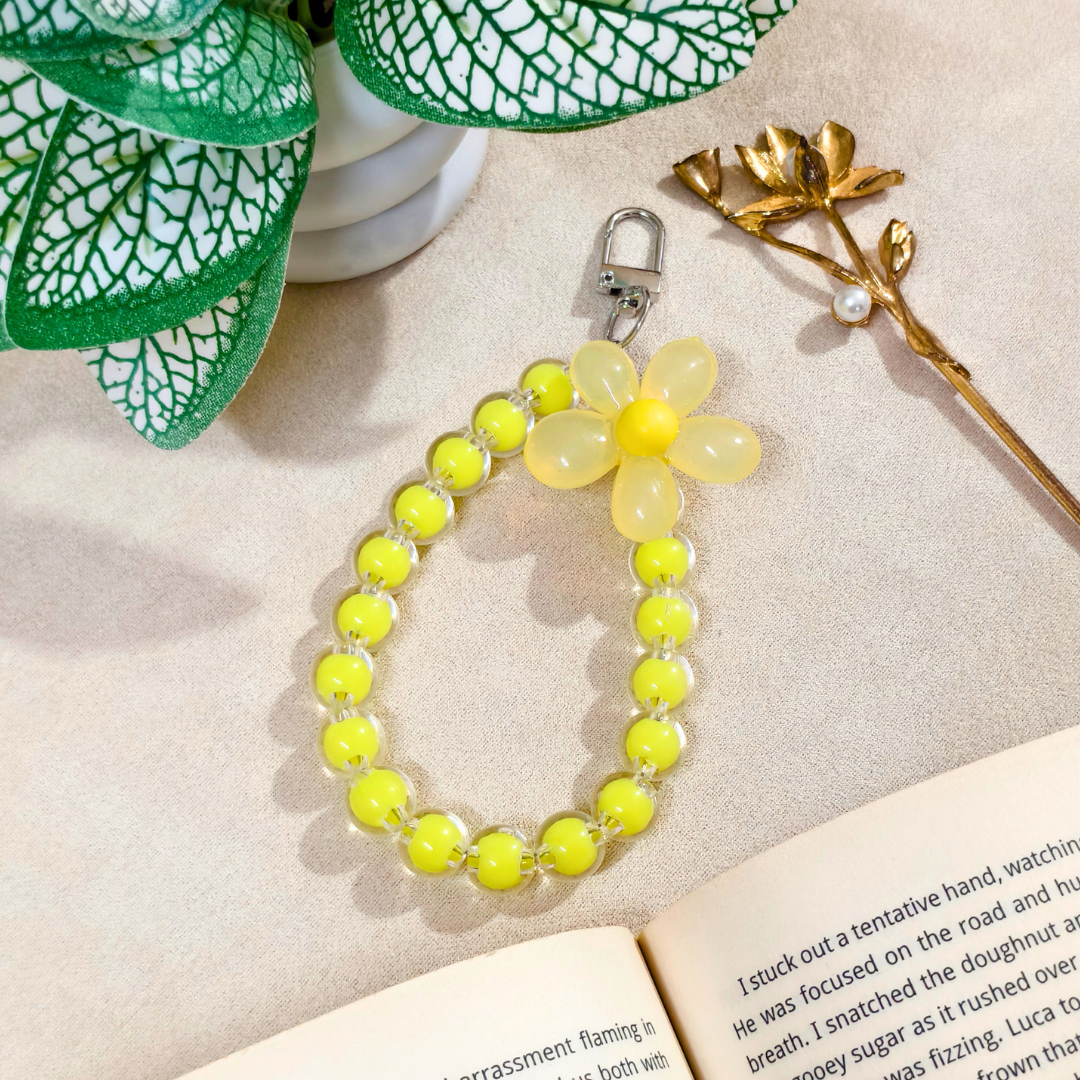 Yellow beaded bracelet with a flower charm on an open book, surrounded by green leaves and a gold branch.