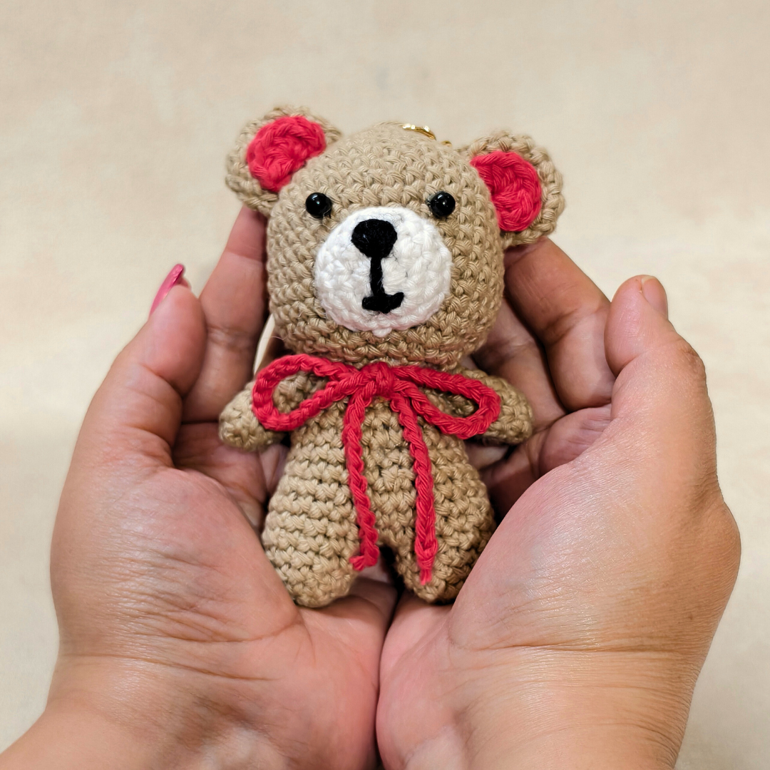 Handmade crochet teddy bear with a red bow held in hands against a plain background