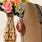 A brown football-shaped keychain with white laces hanging from a silver ring on a tan textured handbag, with a decorative vase of pink flowers in the background.