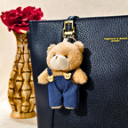 Teddy bear keychain on a black leather bag with a vase of red flowers in the background.