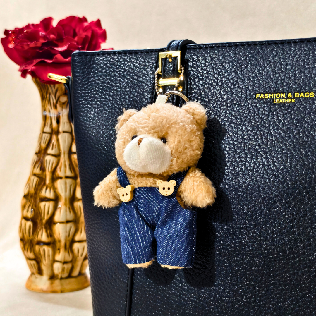 Teddy bear keychain on a black leather bag with a vase of red flowers in the background.