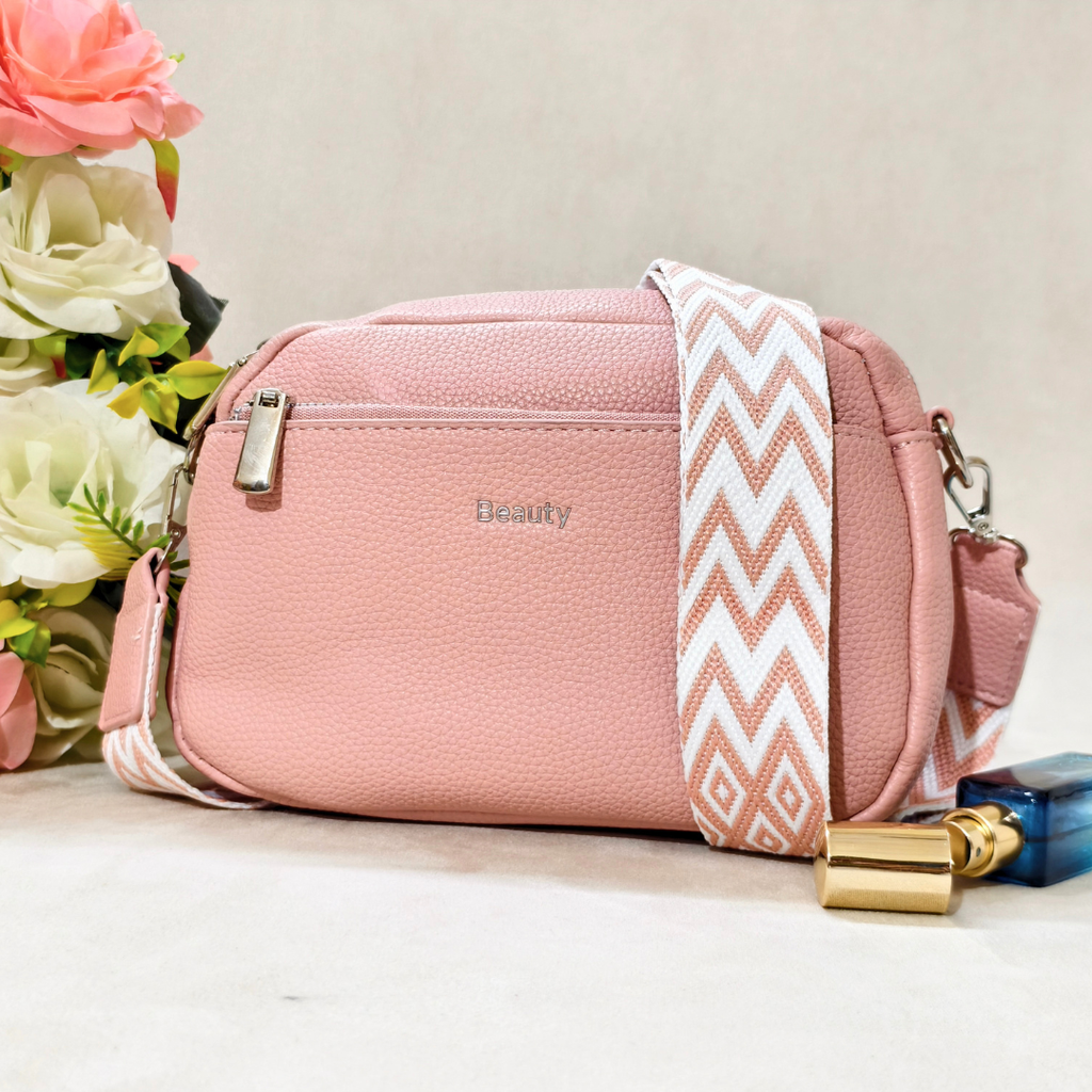 A pink pebbled faux-leather crossbody bag with a wide white and pink chevron-patterned strap, featuring a silver zipper and the word 'Beauty' printed on the front, styled next to a perfume bottle and flowers.