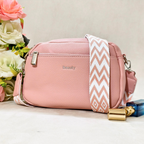 A pink pebbled faux-leather crossbody bag with a wide white and pink chevron-patterned strap, featuring a silver zipper and the word 'Beauty' printed on the front, styled next to a perfume bottle and flowers.