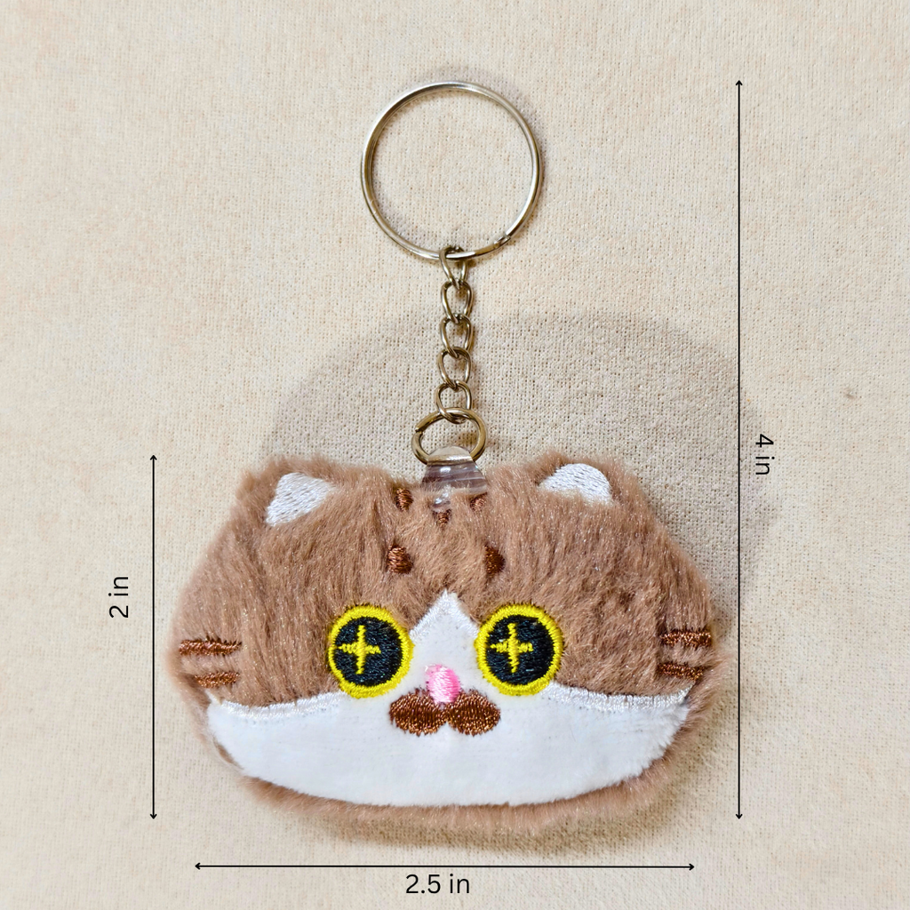 A plush keychain shaped like a small brown and white cat face, held in a person's hand. The keychain has yellow embroidered eyes with black pupils, a pink nose, and a silver metal key ring and chain showing measurements on a beige backgroumd.