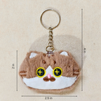 A plush keychain shaped like a small brown and white cat face, held in a person's hand. The keychain has yellow embroidered eyes with black pupils, a pink nose, and a silver metal key ring and chain showing measurements on a beige backgroumd.