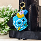 Blue cartoon camera keychain attached to a black bag with a plant in the background