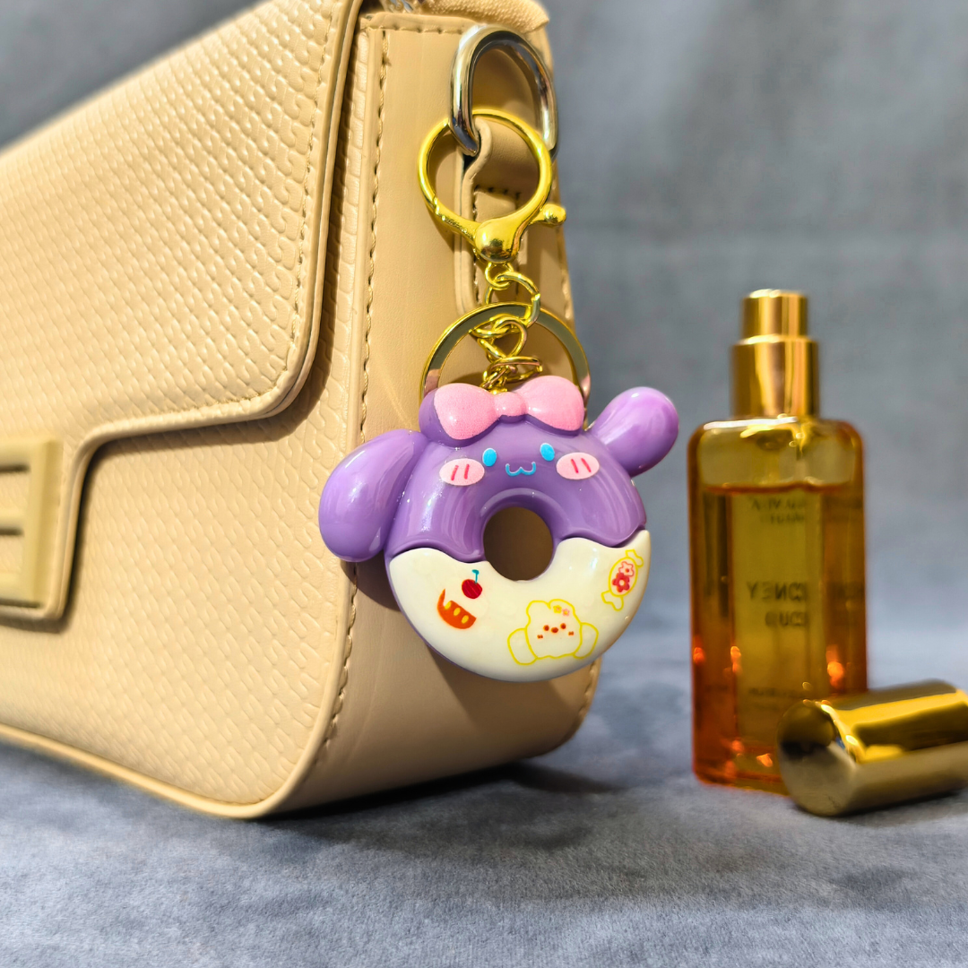 Donut-shaped keychain with a gold chain attached to a beige handbag, next to a gold bottle on a gray surface.