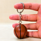 A miniature basketball-shaped keychain held between a person's fingers against a plain beige background.