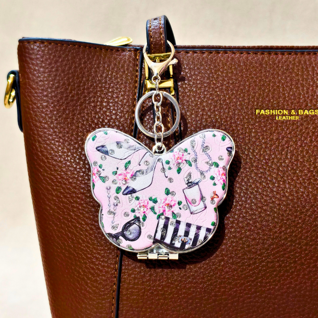 A butterfly-shaped charm keychain with a light pink background, decorated with illustrations of high heels, flowers, and a striped hat, hanging from a brown leather bag.