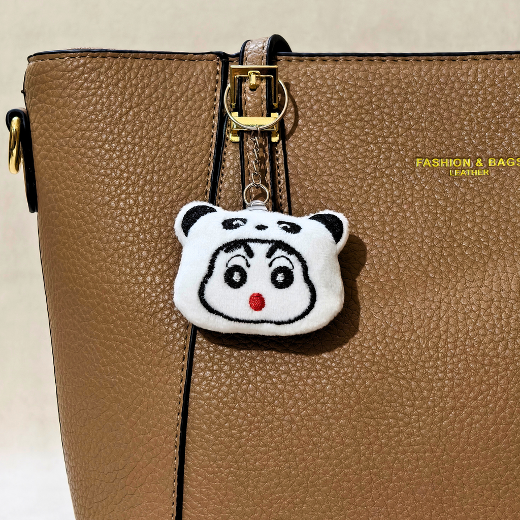 A close-up of a brown textured leather handbag with a gold-tone clasp, featuring a white embroidered panda-face charm with black details and a red nose hanging from the clasp. The bag has gold text on the right side that reads 'FASHION & BAGS PARIS'.