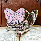 A butterfly-shaped compact mirror with a pink floral and vintage jewelry pattern, resting on an open book with a brown leather bag in the background.