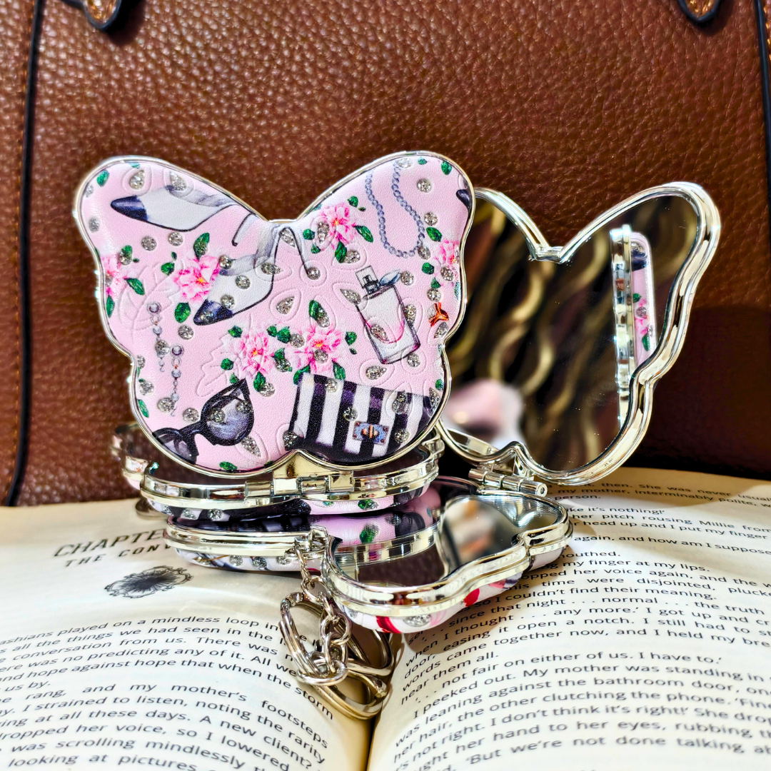 A butterfly-shaped compact mirror with a pink floral and vintage jewelry pattern, resting on an open book with a brown leather bag in the background.