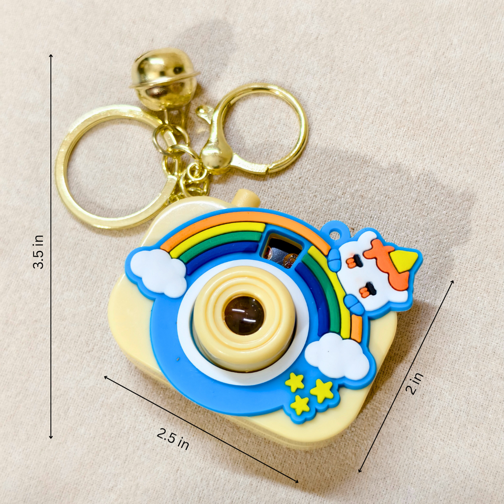 A retro-style camera-shaped keychain with a gold bell and key rings, decorated with a rainbow, clouds, stars, and a cow on a beige fabric background with dimension markings.