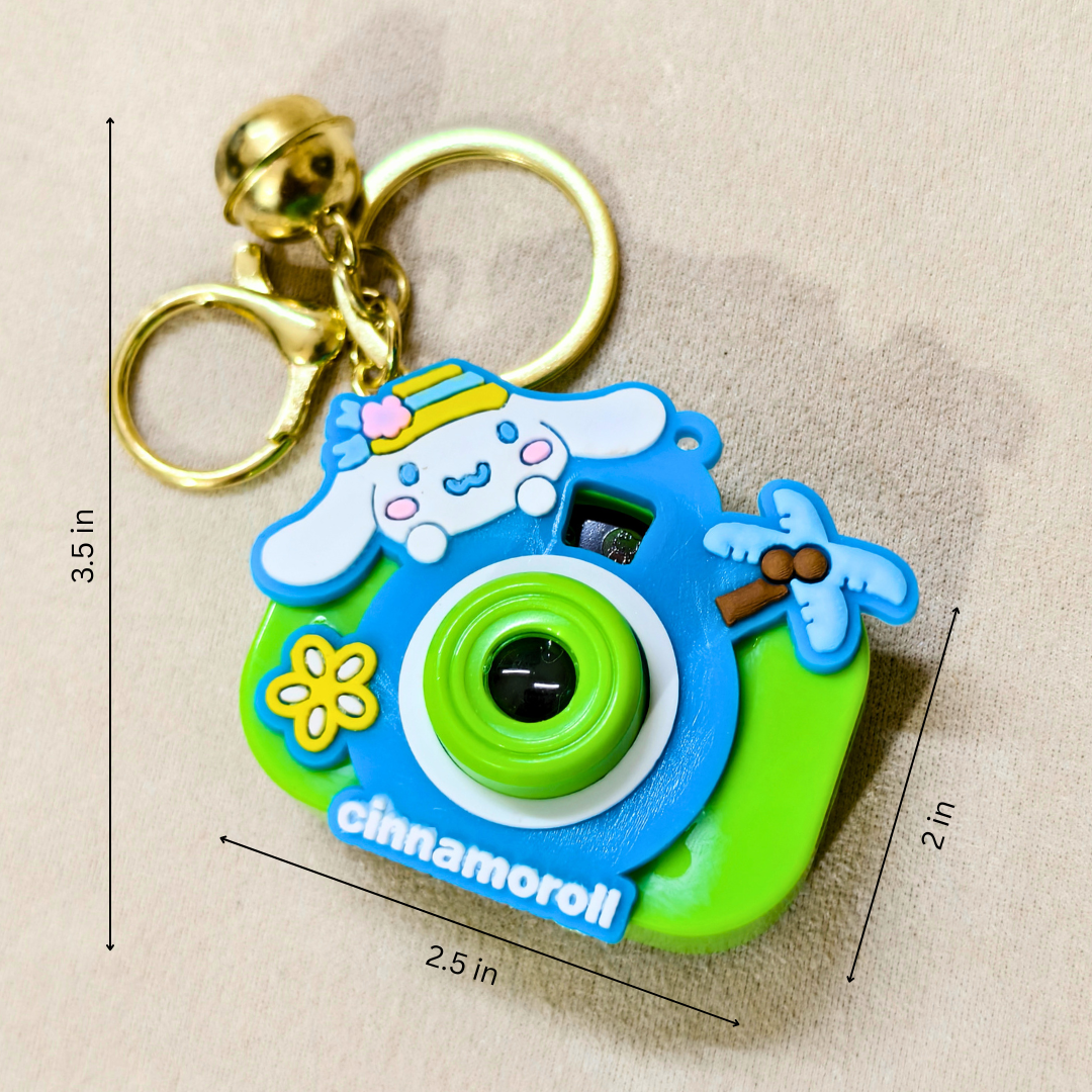 A Cinnamoroll-themed camera-shaped keychain with a gold key ring and clasp, shown on a beige fabric background with printed dimension lines indicating 3.5 in height, 2.