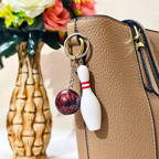 A close-up of a tan textured handbag with a metal key ring holding a bowling-themed charm set: a white bowling pin and a red-and-black marbled bowling ball, shown beside a decorative vase with flowers.
