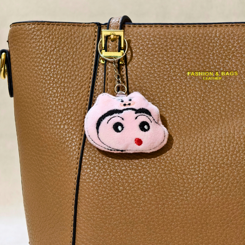 A close-up of a tan pebbled leather handbag with a pink plush character keychain charm hanging from a gold-tone ring and buckle.