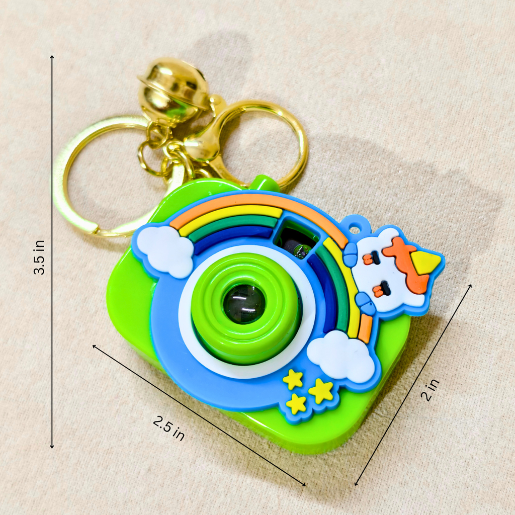 A colorful toy camera keychain with a gold bell and key rings, shown on a beige fabric background with dimension lines indicating 3.5 in height, 2.