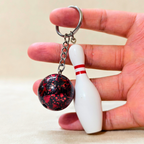 A hand holding a miniature bowling-themed keychain with a white bowling pin charm and a red and black marbled ball on a silver split ring, set against a neutral beige background.