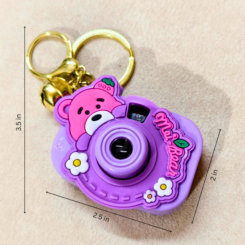 A purple and pink cartoon camera keychain with a gold key ring, shown on a beige fabric background with dimension lines indicating 3.5 in height, 2.