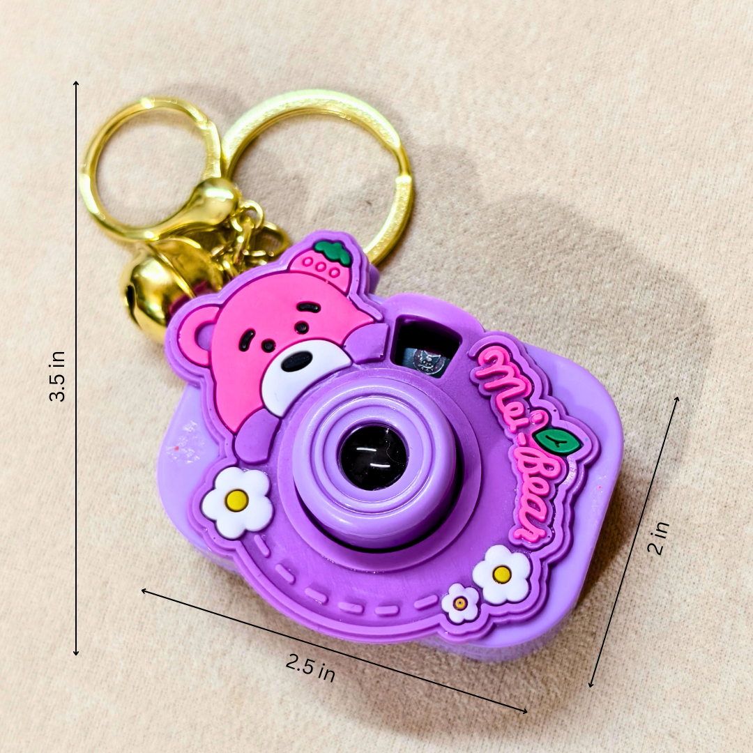 A purple and pink cartoon camera keychain with a gold key ring, shown on a beige fabric background with dimension lines indicating 3.5 in height, 2.
